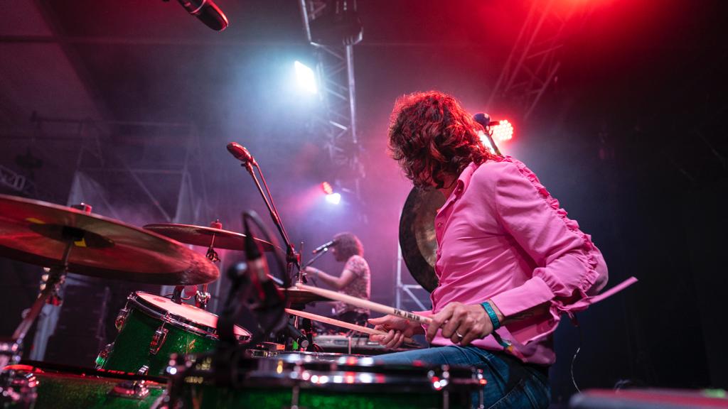 Artista toca la batería mirando a su compañero, de espaldas a la cámara.